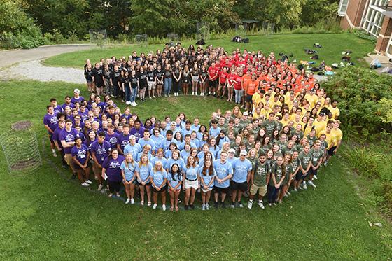 Photo from above, showing a large group of Chatham University students in multicolored T-shirts stand in a C shape together outside on green grass. 